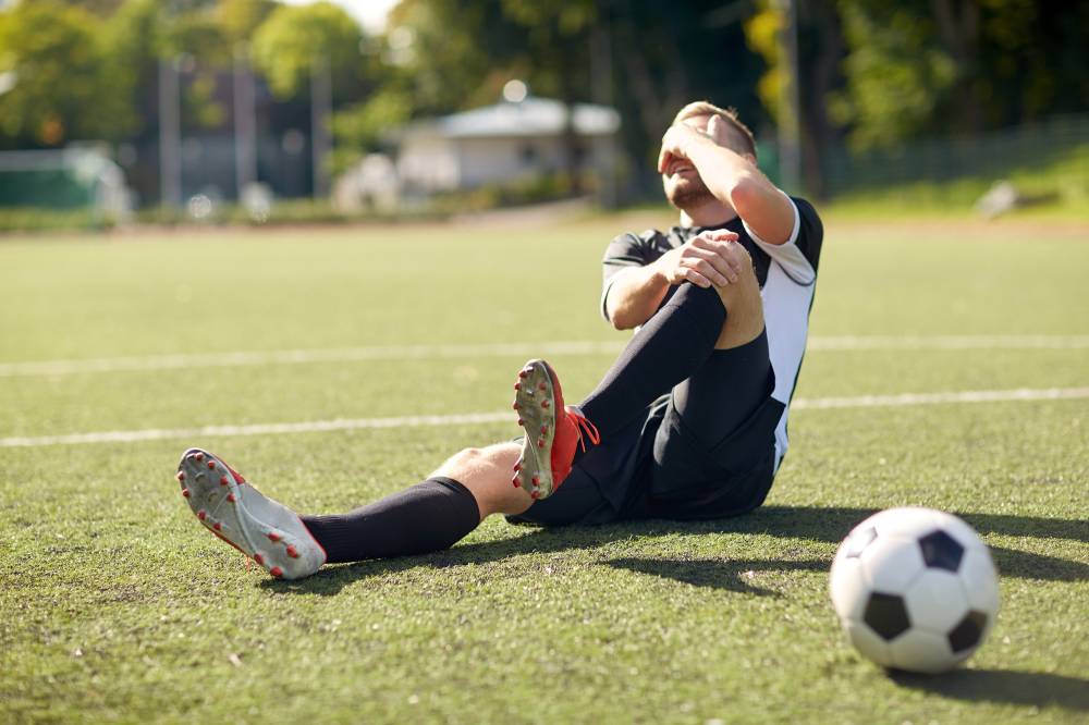 Injured soccer player sitting on the grass while he grasps his knee in pain and covers his face.